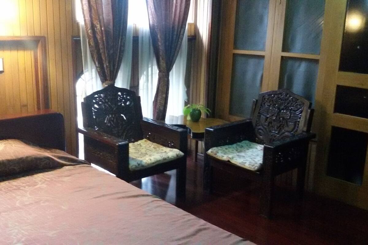 A room with a bed, two wooden chairs with cushioned seats, a small table with a plant, and a window with curtains.