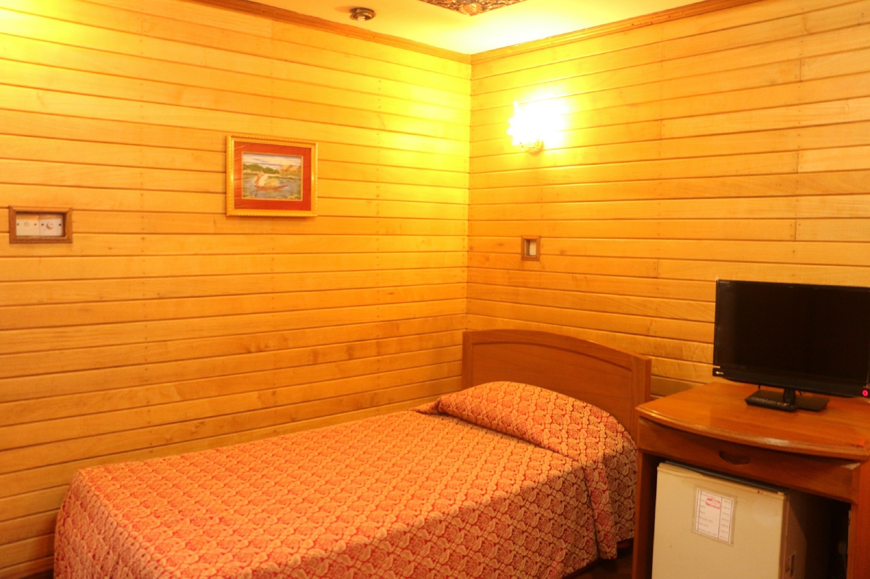A small, cozy room with wooden walls, a single bed with a patterned bedspread, a television on a wooden stand, and a small refrigerator.