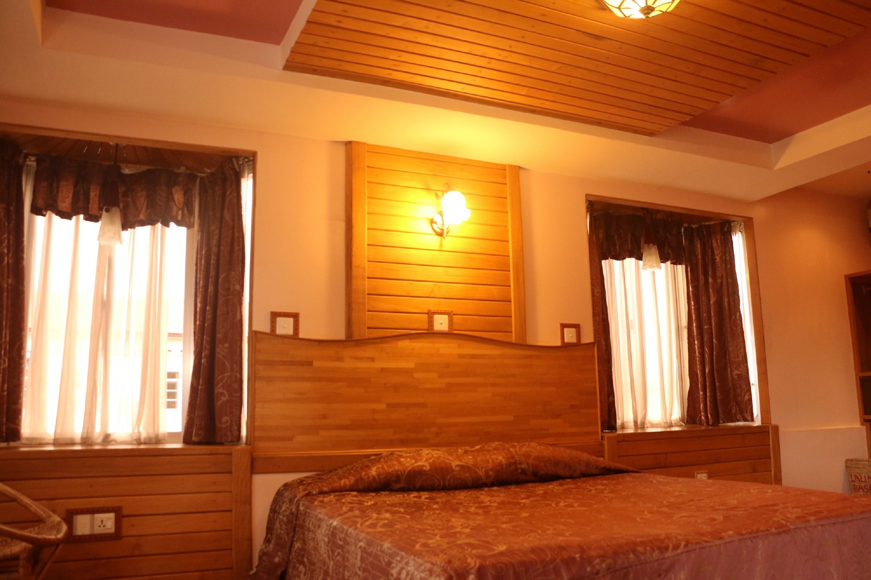 A room with a wooden headboard, a bed with a patterned bedspread, and two windows with curtains.