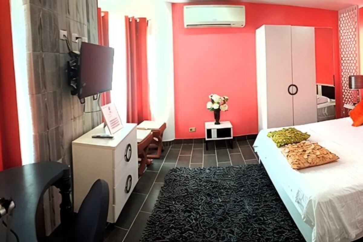 A bedroom with a bed, a desk, a chair, a TV, and an air conditioning unit.