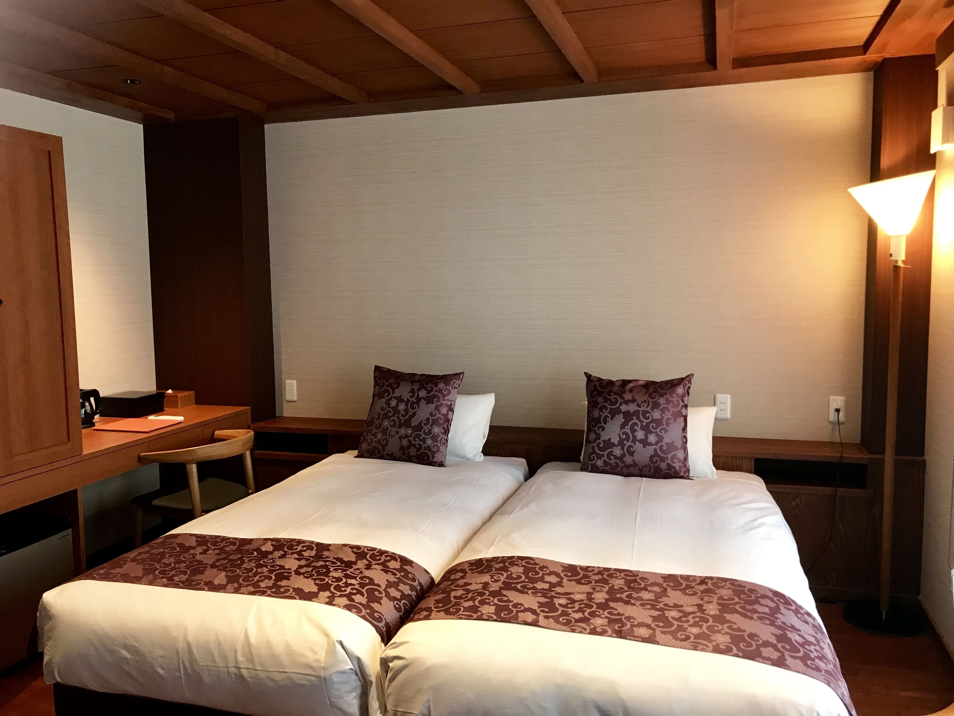 A hotel room with two beds, a wooden desk, and a nightstand with a lamp.
