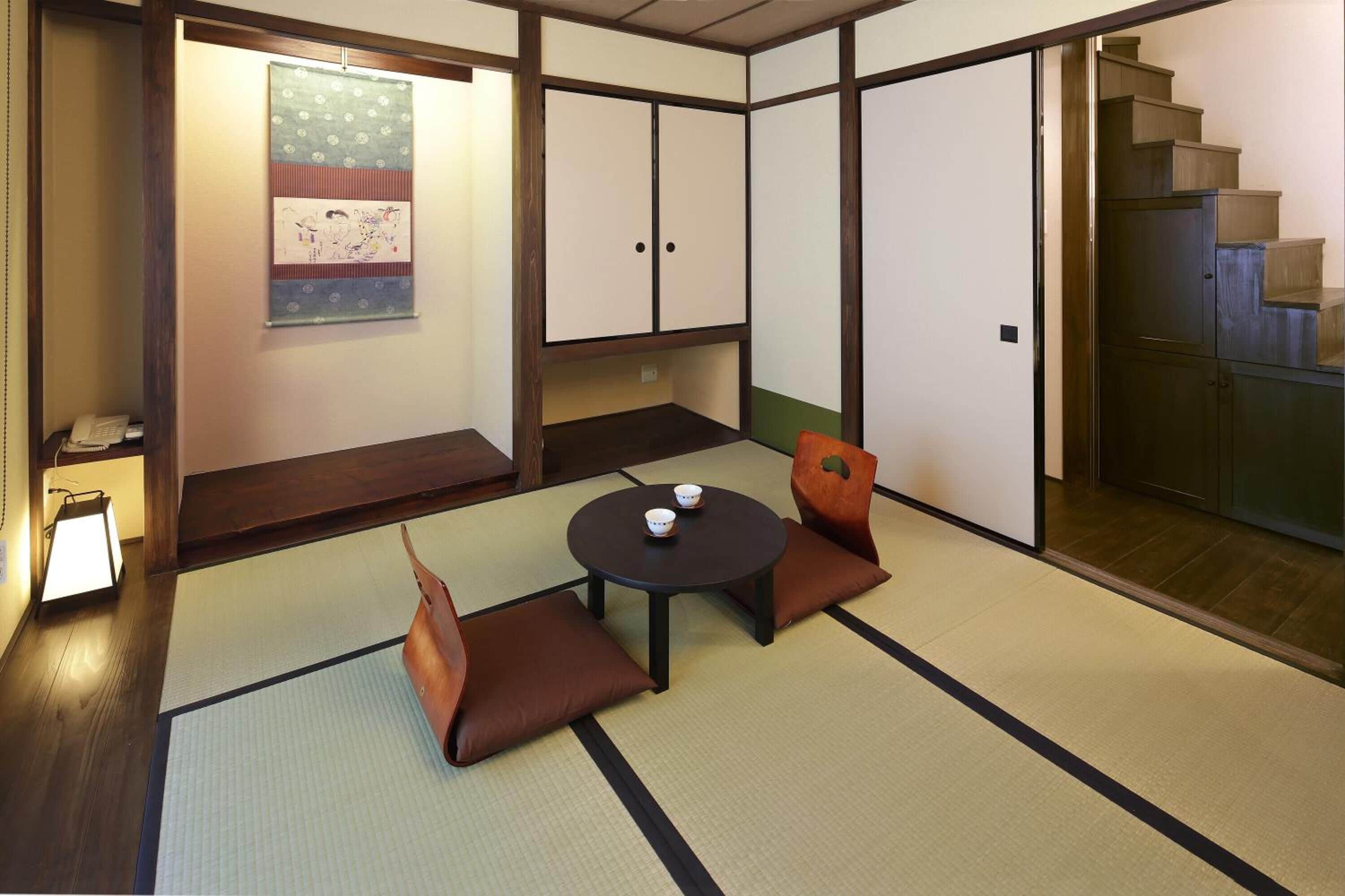 Una tradizionale stanza giapponese con pavimento in tatami, un tavolino con cuscini, un'opera d'arte appesa e una scala.