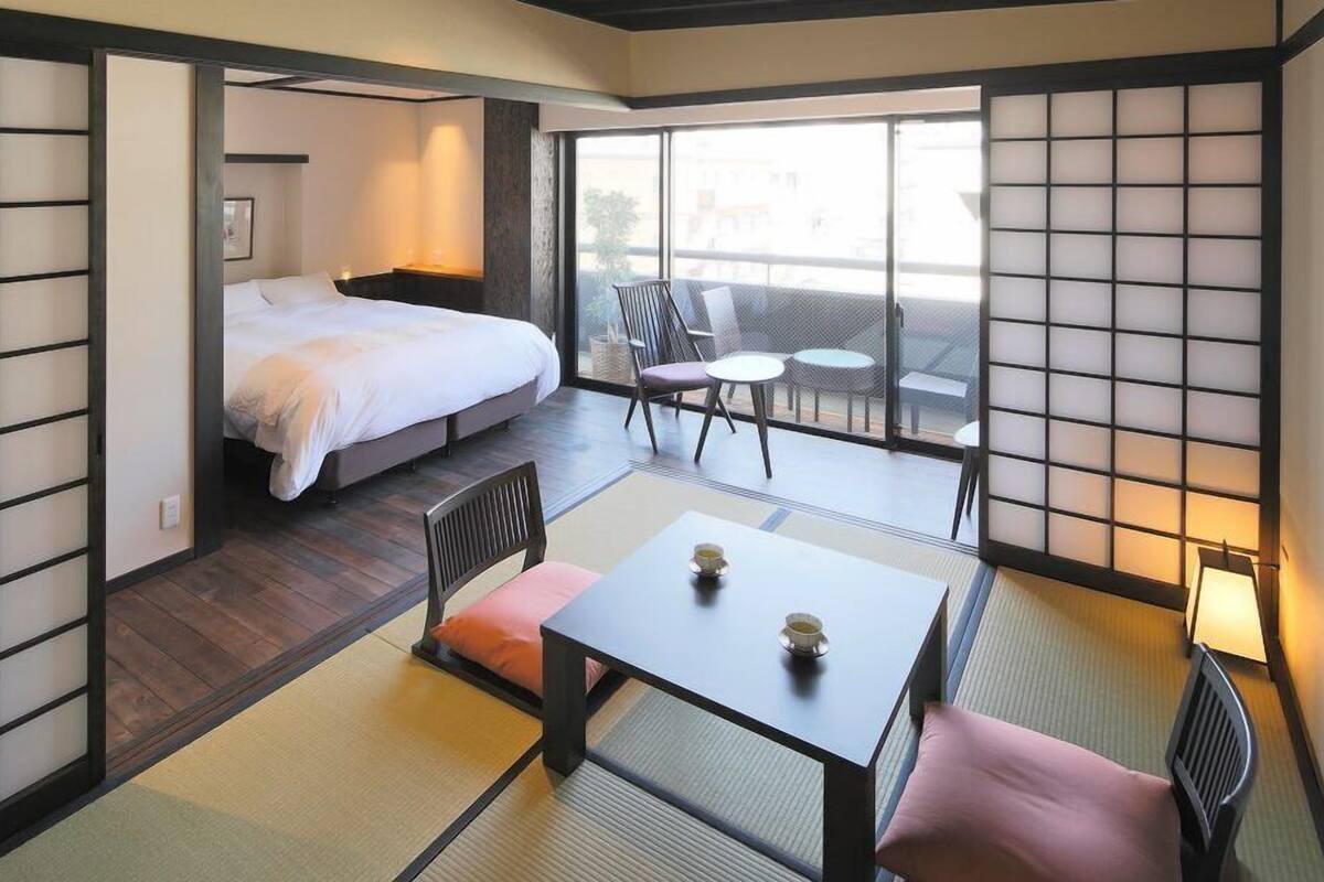 Una camera d'albergo con un letto, un tavolino con una tazza, una sedia e un balcone con vista.