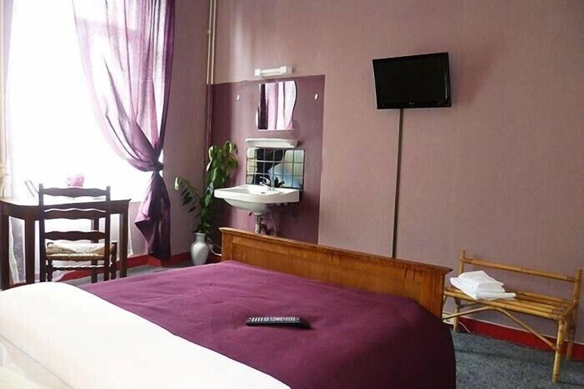 A hotel room with a bed, a wooden chair, a small table, a TV mounted on the wall, and a bathroom with a sink and mirror.