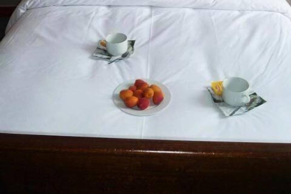 A neatly made bed with white linens, a wooden headboard, and a statue of an angel on the bed. There are two cups and a plate of fruit on the bed.