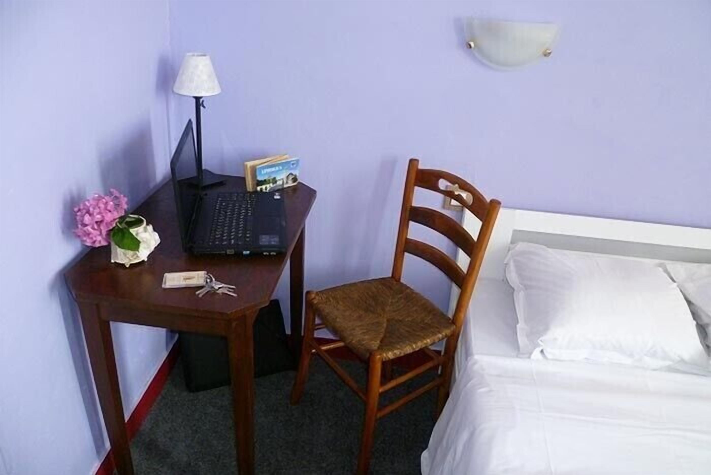 A hotel room with a bed, a wooden chair, a desk with a laptop, a lamp, and a vase with flowers.