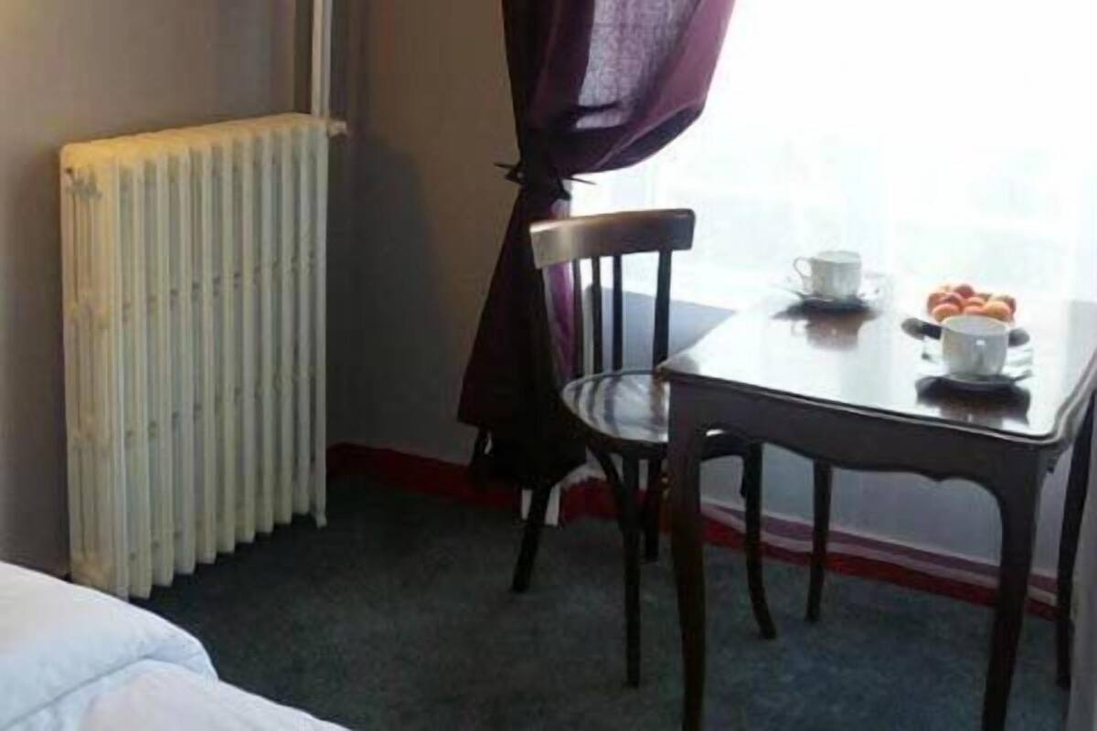 A hotel room with a bed, a table with a cup and pastries, a chair, a radiator, and a window with purple curtains.