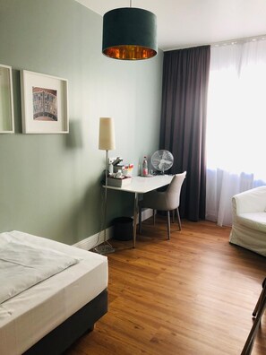A hotel room with a bed, a desk with a chair, a standing fan, and a large window with curtains. - guestified. AYUNA Hotel (Düsseldorf)