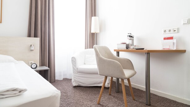 Habitación de hotel con cama, una silla, escritorio con lámpara, cafetera y una mesita con taza y caja.