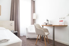 A hotel room with a bed, a chair, a desk with a lamp, a coffee maker, and a small table with a cup and a box. - guestified. AYUNA Hotel (Düsseldorf)