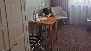 Habitación de hotel con cama, escritorio con silla, una mesita, un cesto de basura y ventana con cortinas.