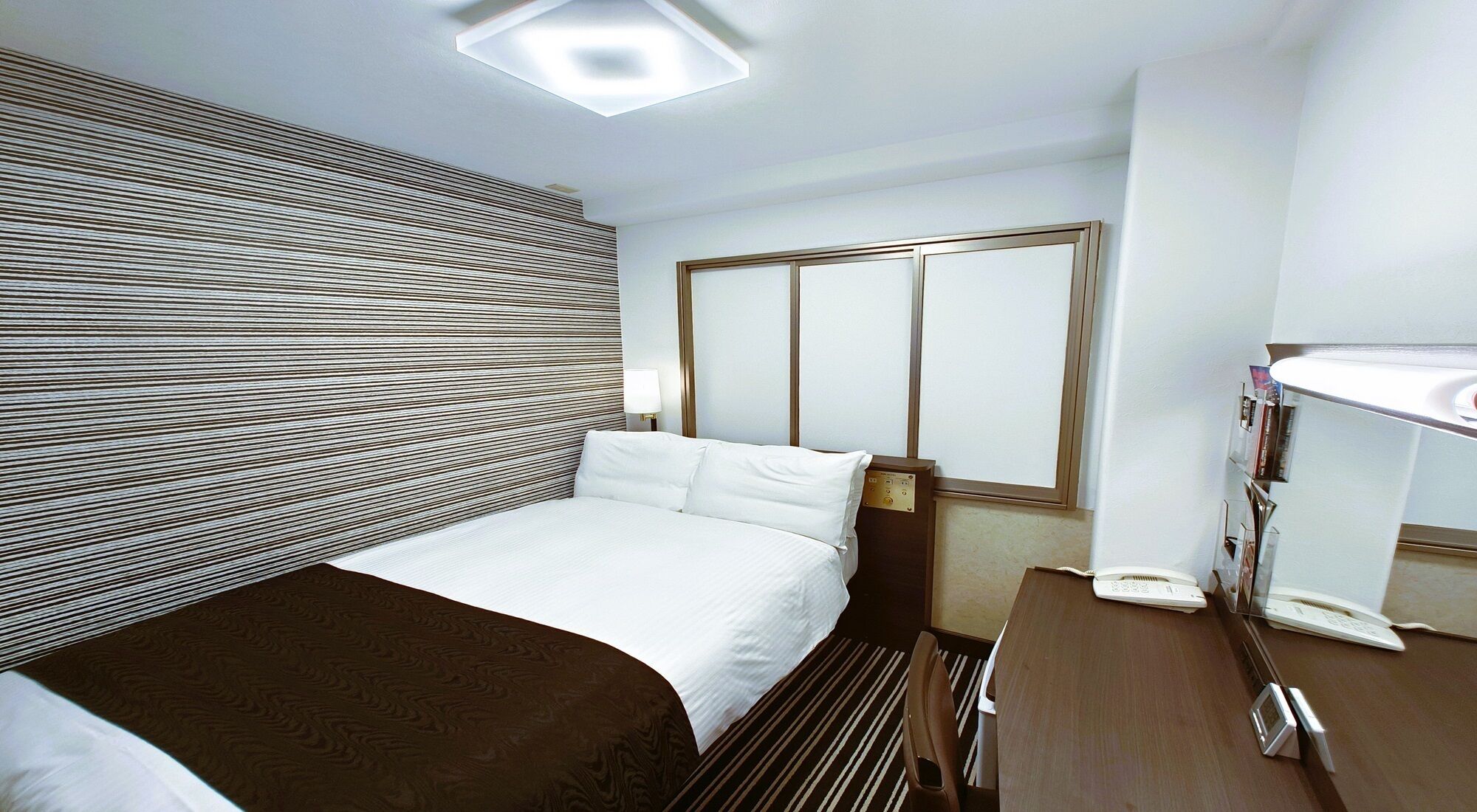 A hotel room with a bed, bedside table, lamp, and a window with blinds.