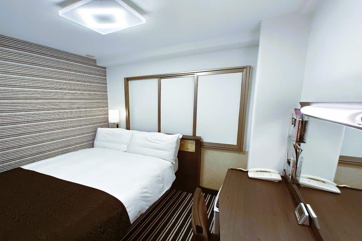 A hotel room with a bed, bedside table, lamp, and a window with blinds.