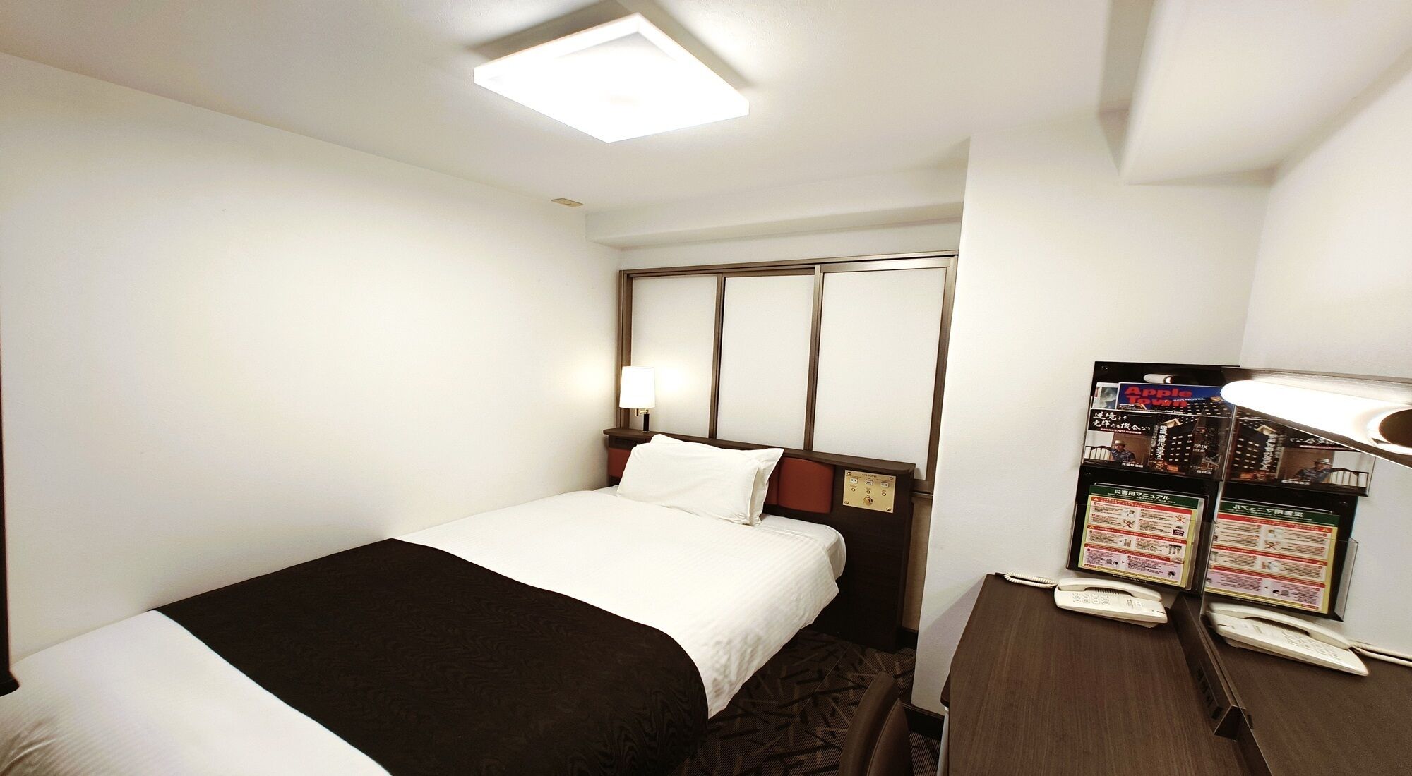 A hotel room with a bed, a desk with a telephone, and a shelf with informational brochures.