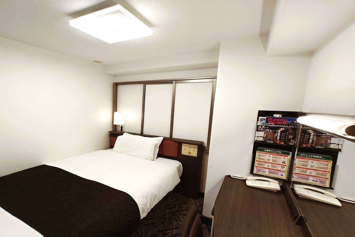 A hotel room with a bed, a desk with a telephone, and a shelf with informational brochures.