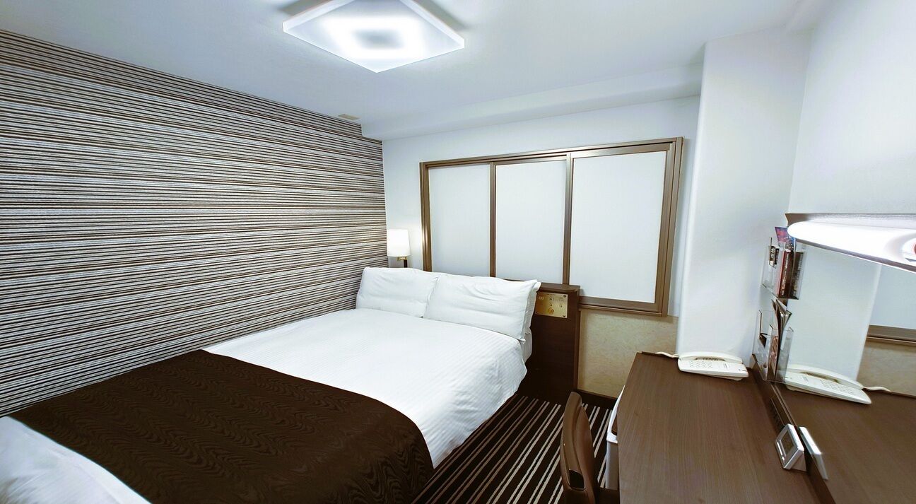 Una habitación de hotel con cama, mesita de noche, lámpara y ventana con persianas.