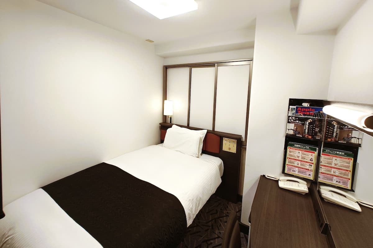 A hotel room with a bed, bedside table, lamp, and a bulletin board with informational posters.
