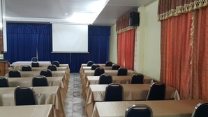 Un salón de conferencias con filas de mesas y sillas, una pantalla de proyector y cortinas.