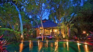 Un edificio de estilo tradicional con tejado de baldosas, rodeado de exuberante vegetación y una piscina con luces que se reflejan en el agua.