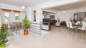 A spacious living area with a dining table, chairs, a sofa, and a TV. There are potted plants and a staircase with a railing. - Diogenis Home (Rethymno)