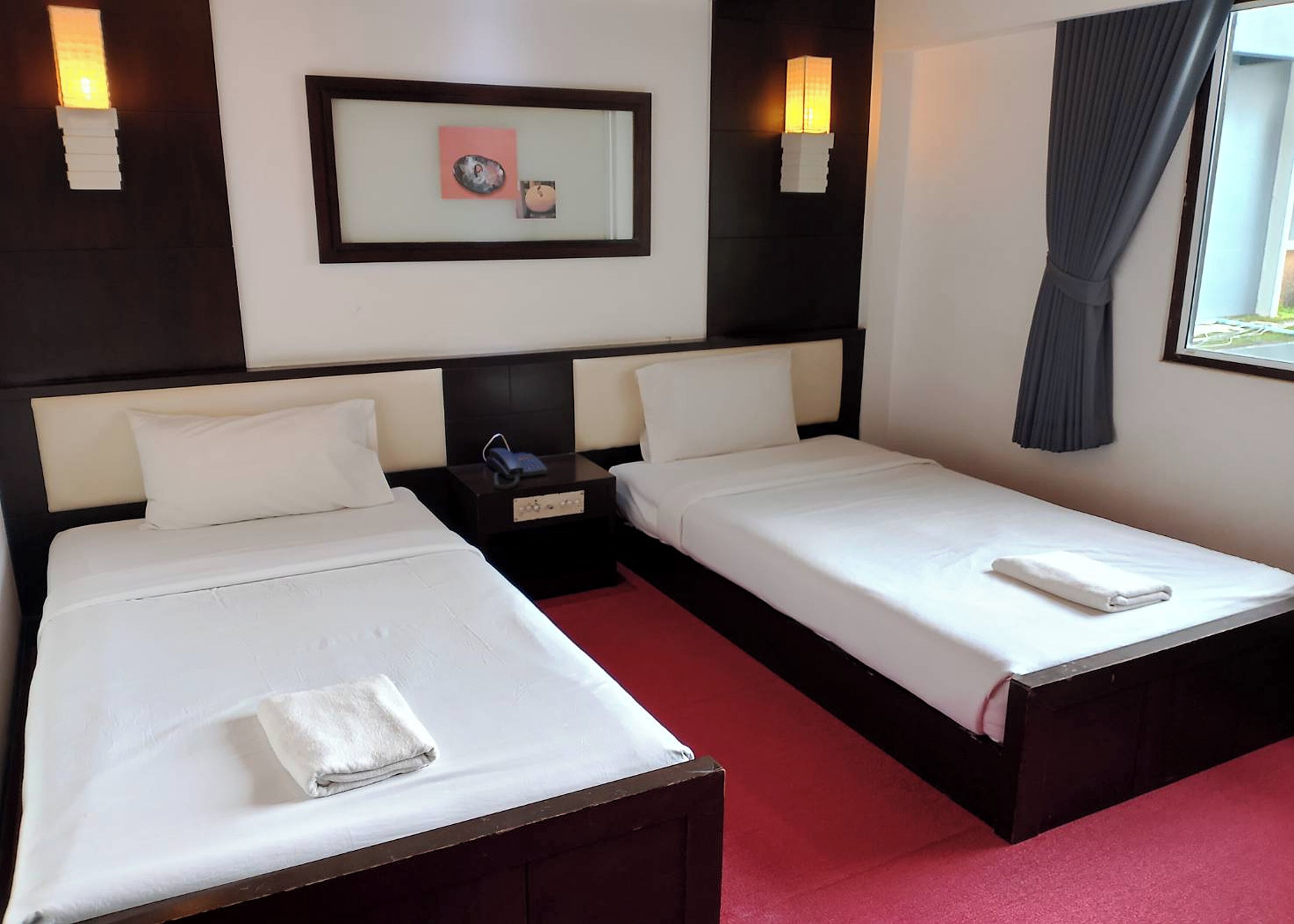 A hotel room with two single beds, a nightstand with a phone, a framed picture on the wall, and a window with curtains.