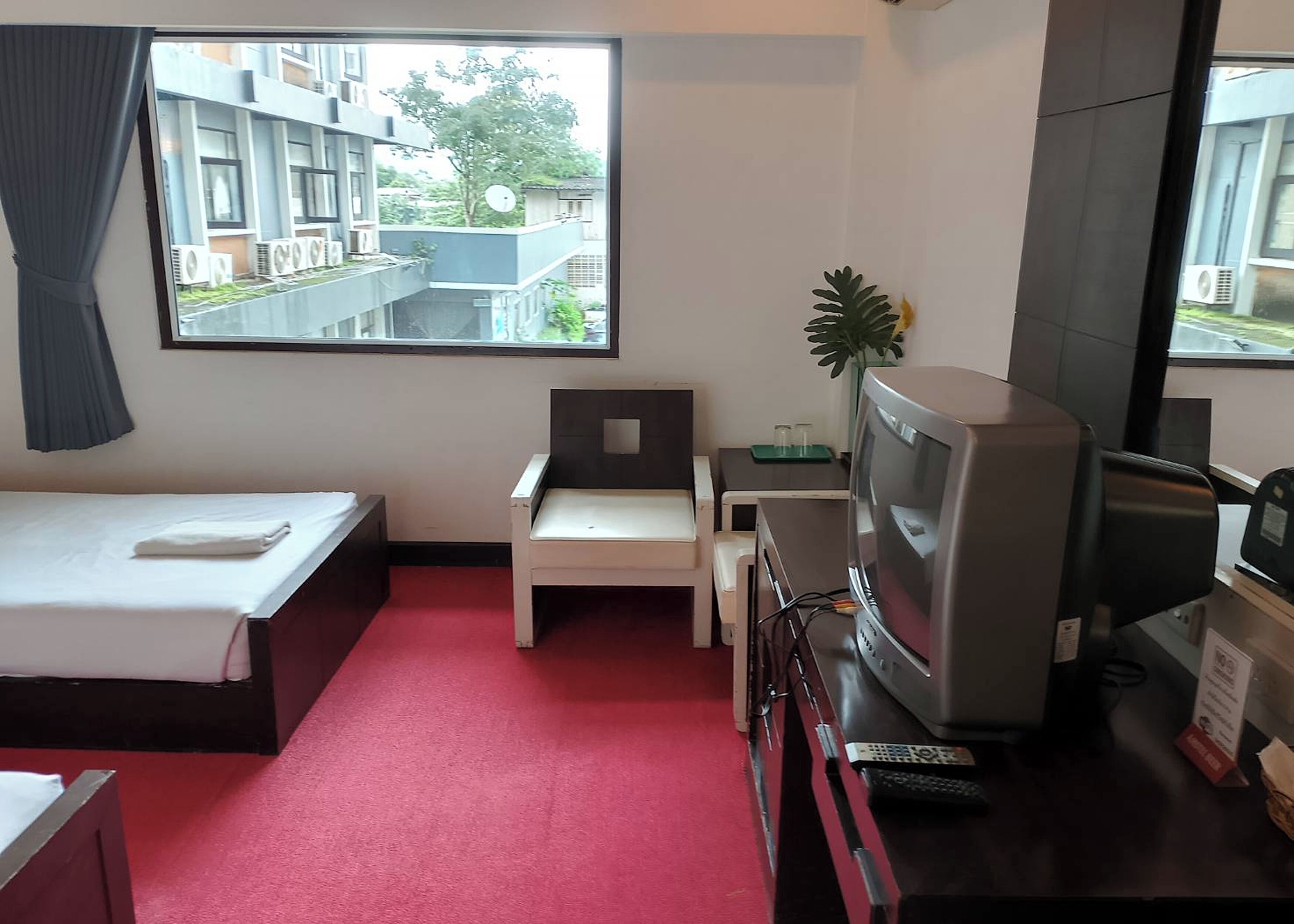 A hotel room with a bed, a desk, a television, and a window with a view of buildings and greenery.