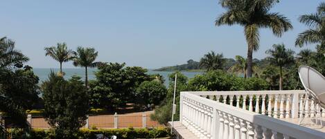 Un balcon doté d’une balustrade blanche, donnant sur un jardin planté de palmiers et offrant une vue sur la mer.