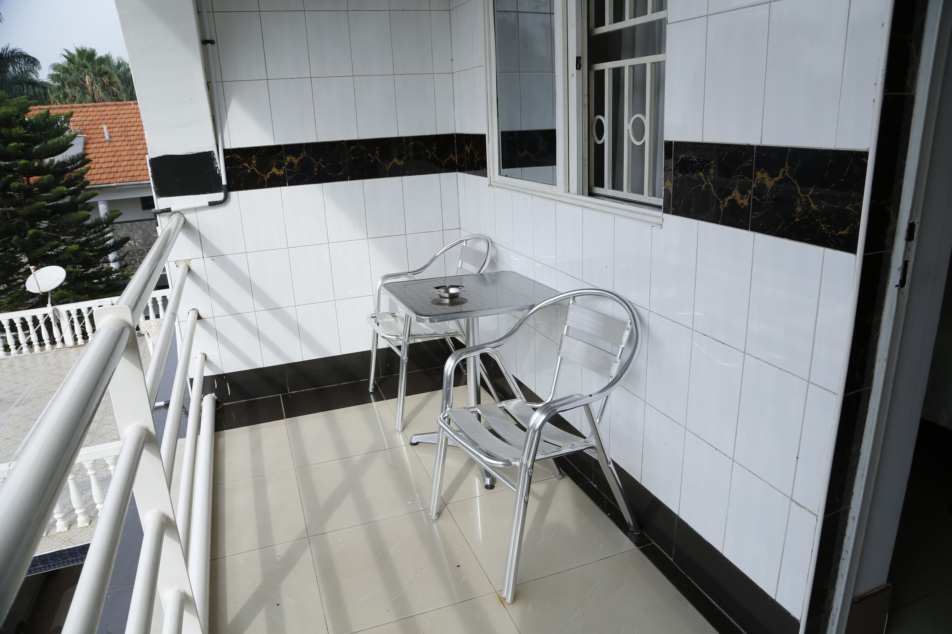 Un balcón con una mesa metálica y dos sillas, paredes de baldosas blancas con detalles negros, y una ventana con rejas de metal.