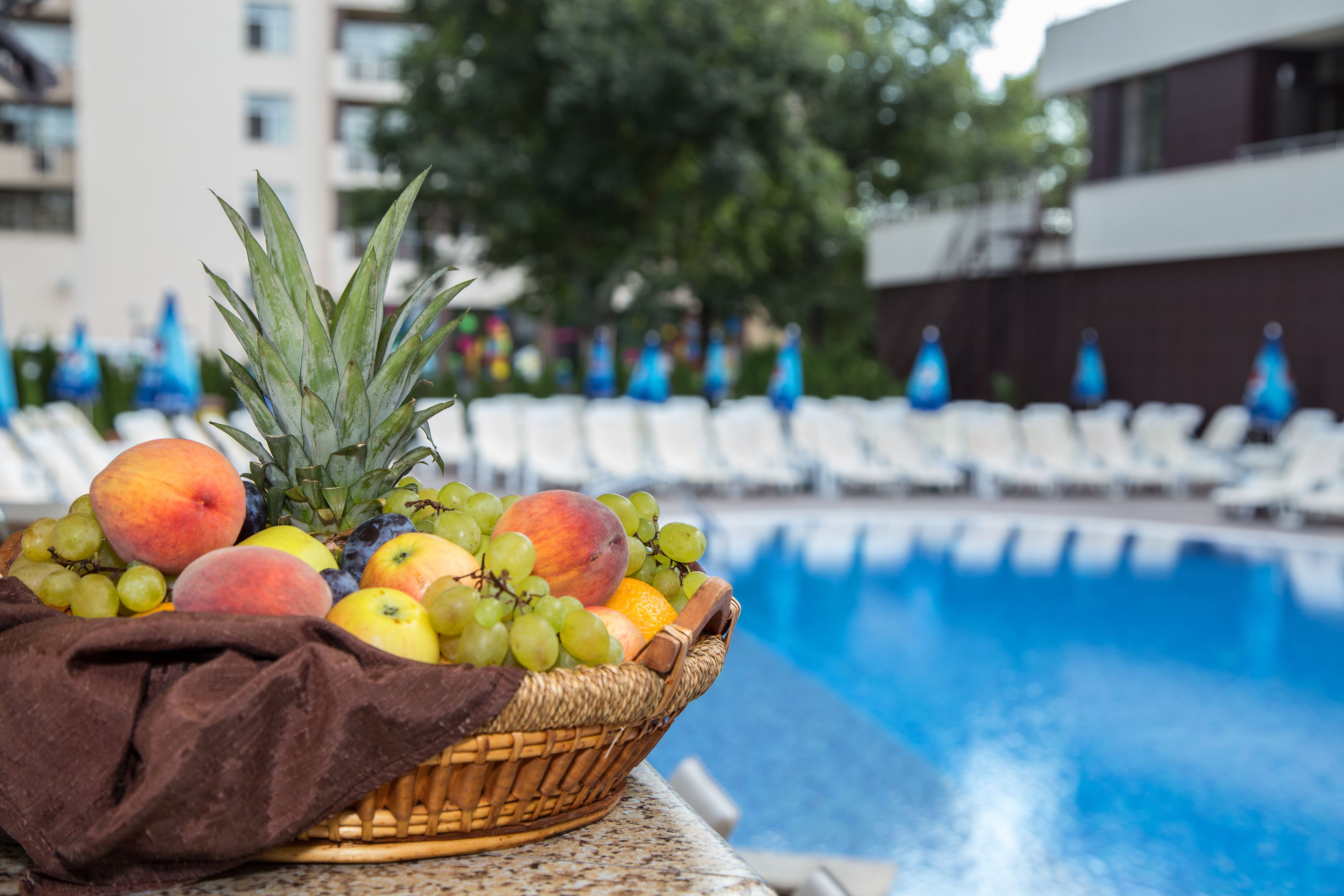 A fruit basket on a poolside table with a pool and lounge chairs in the background.