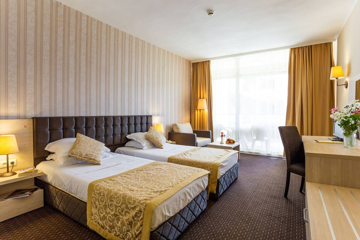 A hotel room with two beds, a desk, a chair, a lamp, and a window with curtains.