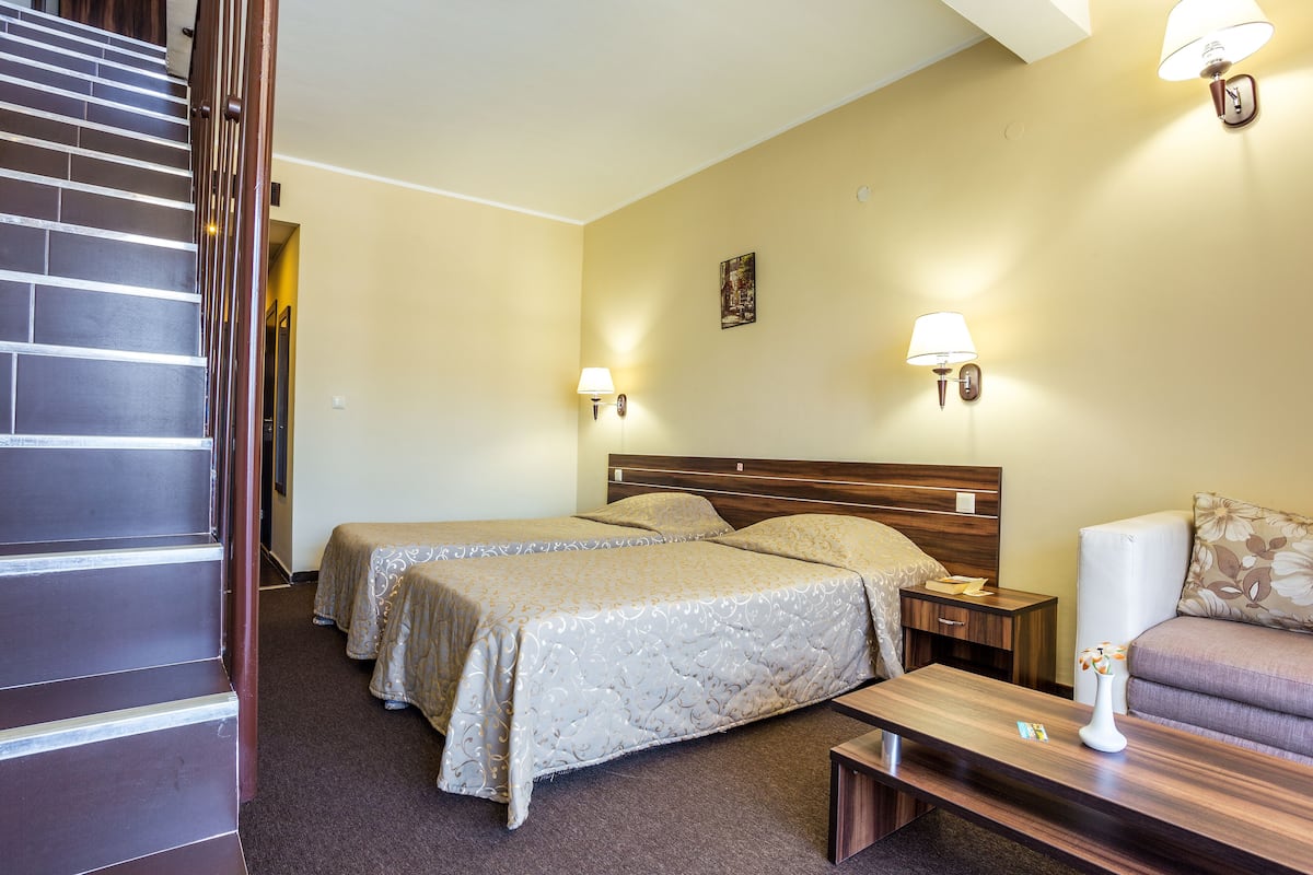 Une chambre d’hôtel avec deux lits, une tête de lit en bois, des tables de chevet, une petite table, un canapé et une lampe.