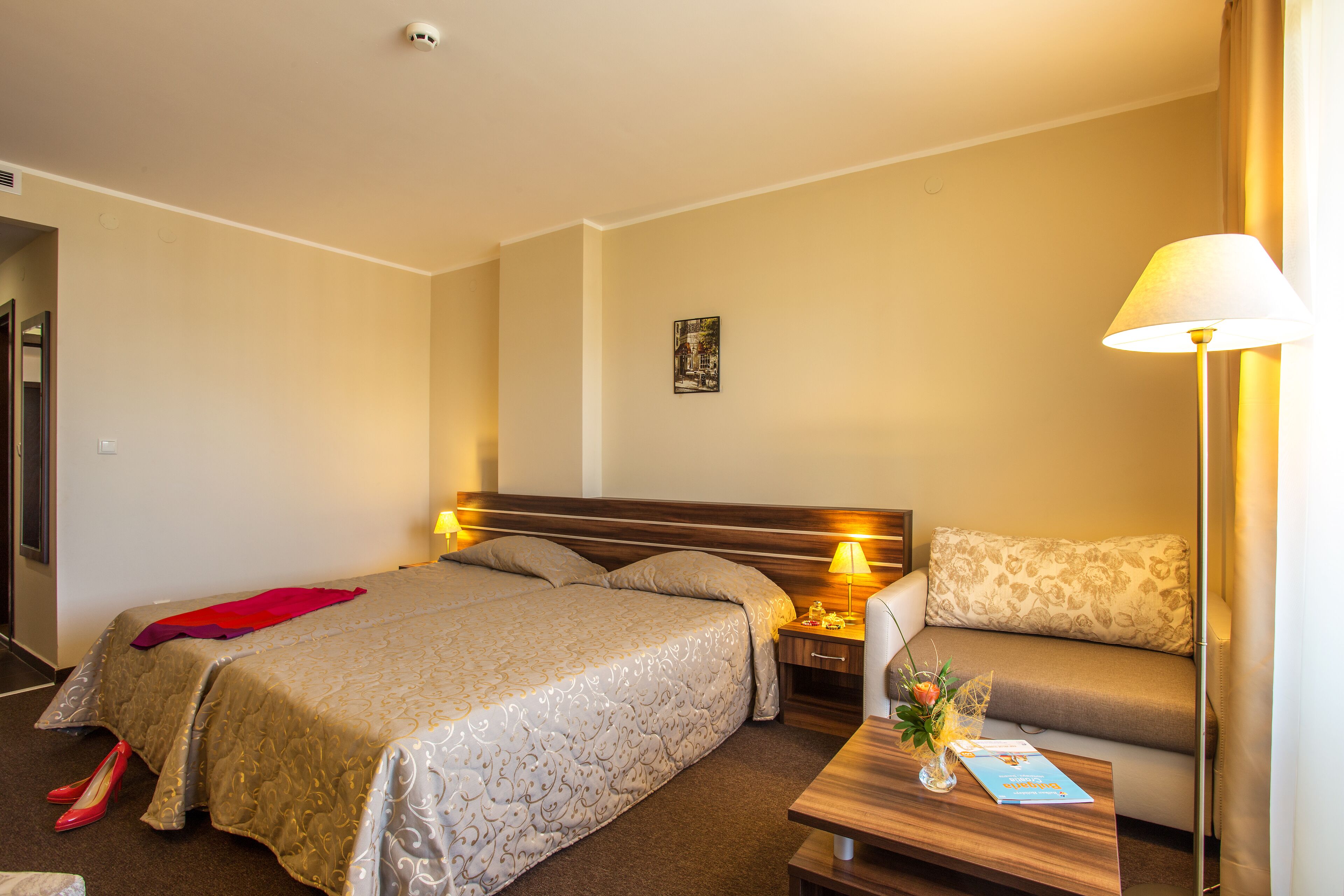 Hotelkamer met een bed, een bank, een nachtkastje, een lamp en een klein tafeltje met een boek en bloemen.