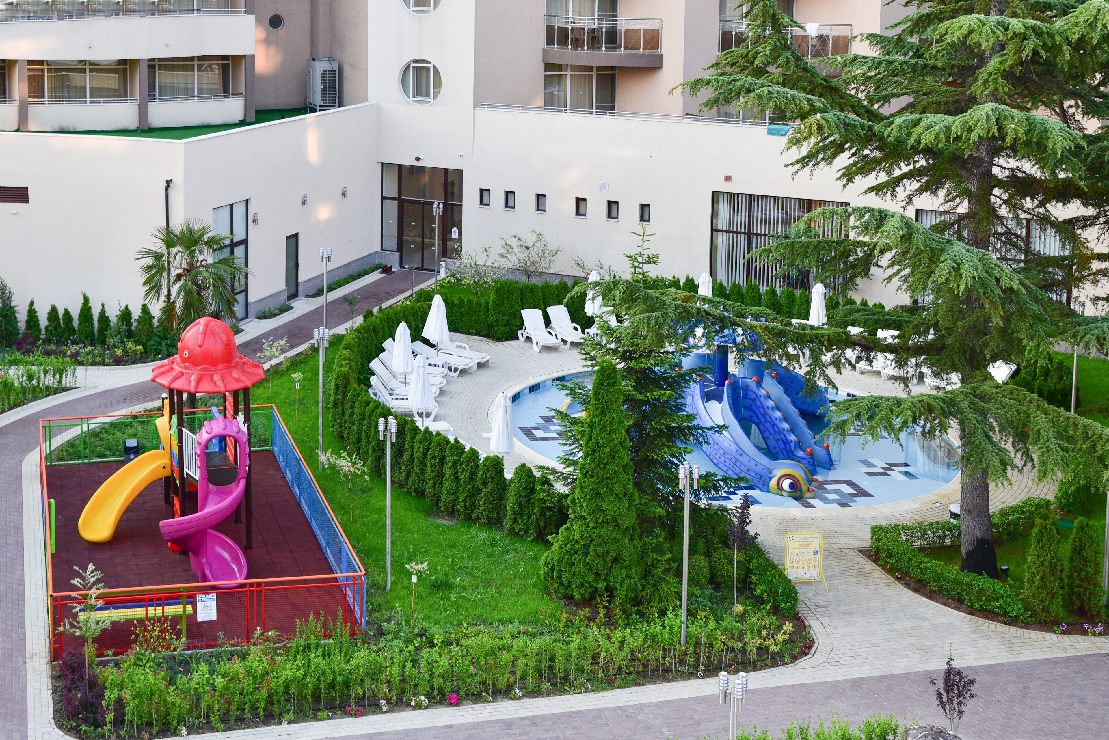 Un terrain de jeux avec des toboggans et un espace de jeux aquatiques, entouré de verdure et bordé d’un bâtiment doté de balcons.