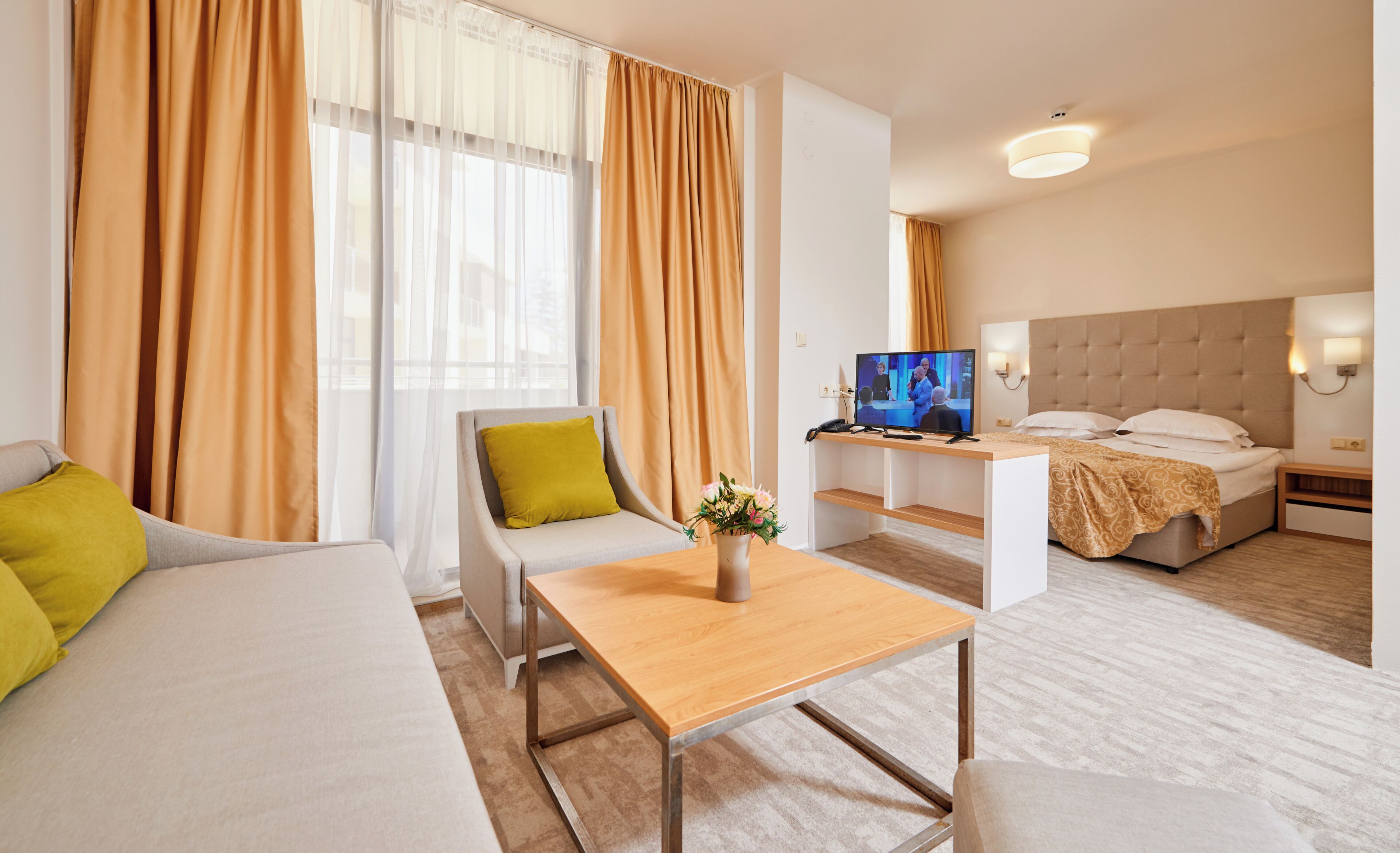 A modern hotel room with a sofa, a small table with a vase, a bed, a television, and a nightstand.