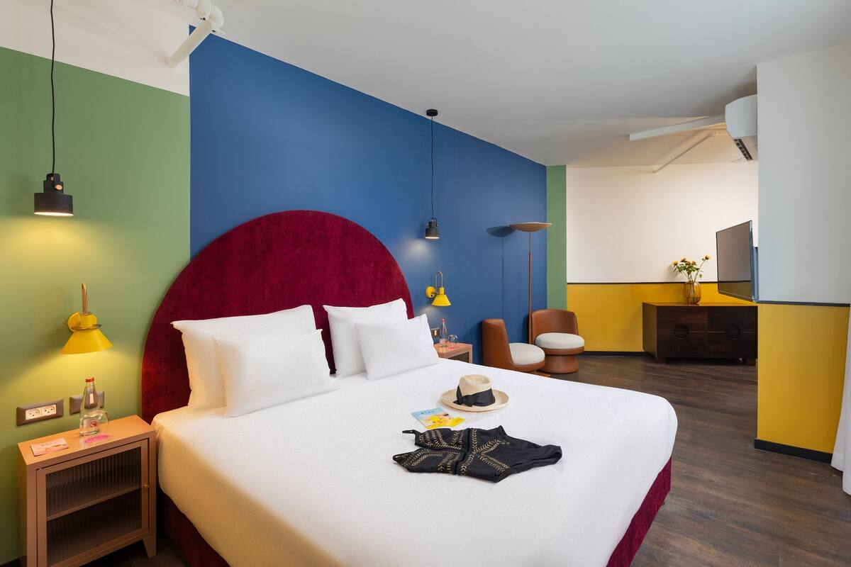 Una habitación de hotel con una cama grande, cabecera roja, mesita de noche con lámpara, una silla, una pequeña mesa con un florero y un televisor.