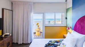 Hotelkamer met een bed, een raam met uitzicht op zee, een klein balkon met een tafel en zonnebloemen, en een houten dressoir.