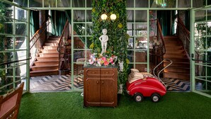 Een trap met een houten leuning, een groene wand begroeid met klimplanten, een beeld en een rode speelgoedauto.
