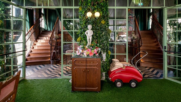Een trap met een houten leuning, een groene wand begroeid met klimplanten, een beeld en een rode speelgoedauto.