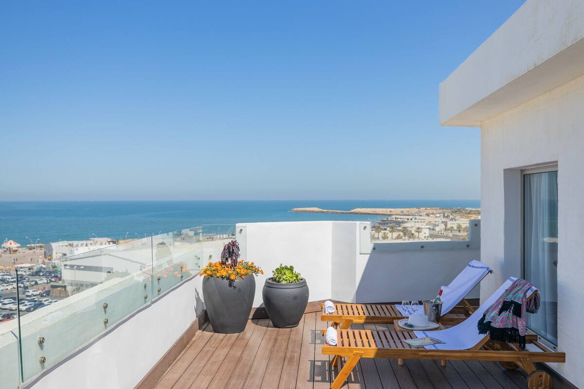 Un balcón con dos macetas grandes, una silla de lounge de madera y vista al mar.