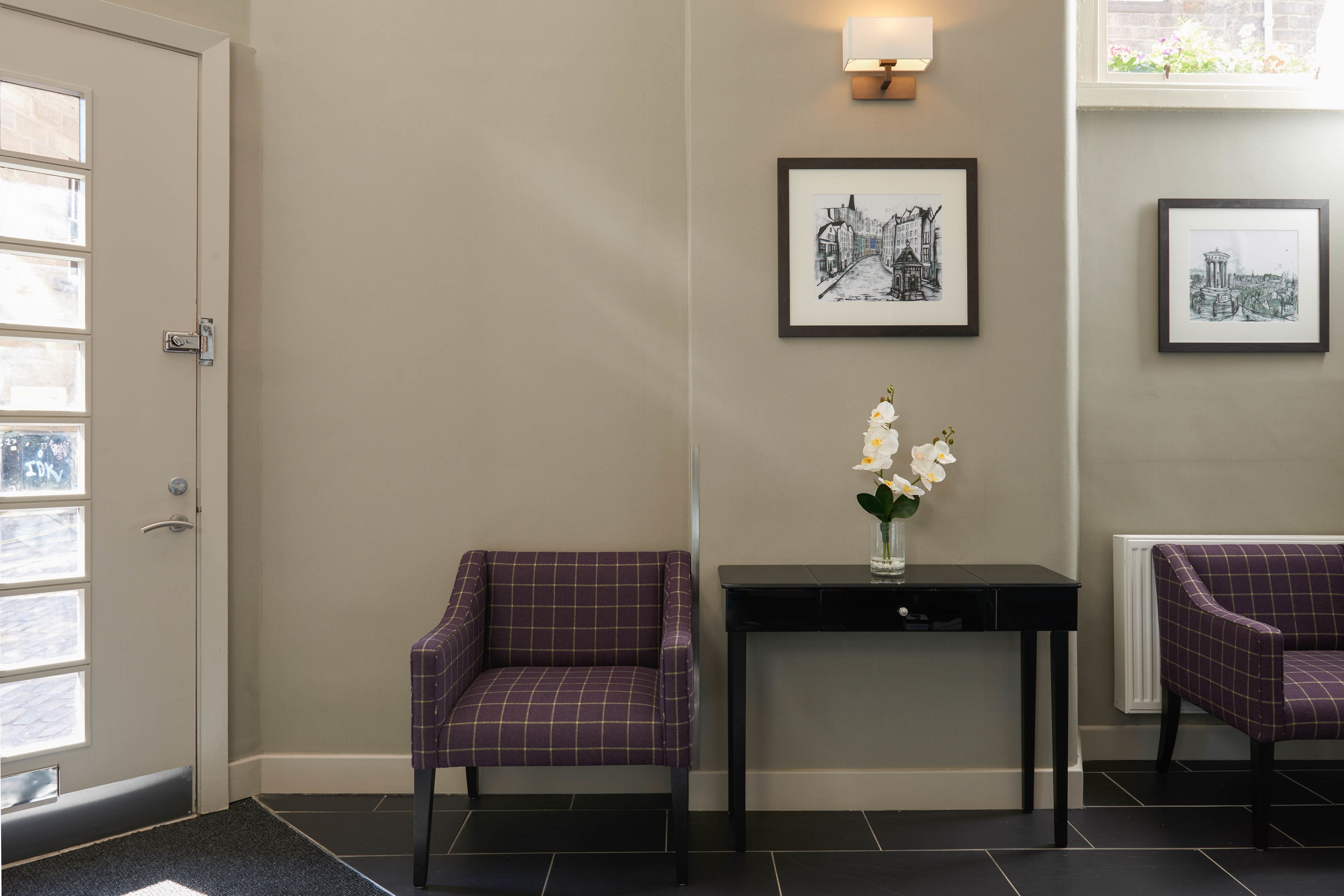 A reception area with two purple armchairs, a black console table with a vase of white flowers, and framed black and white cityscape pictures on the wall.