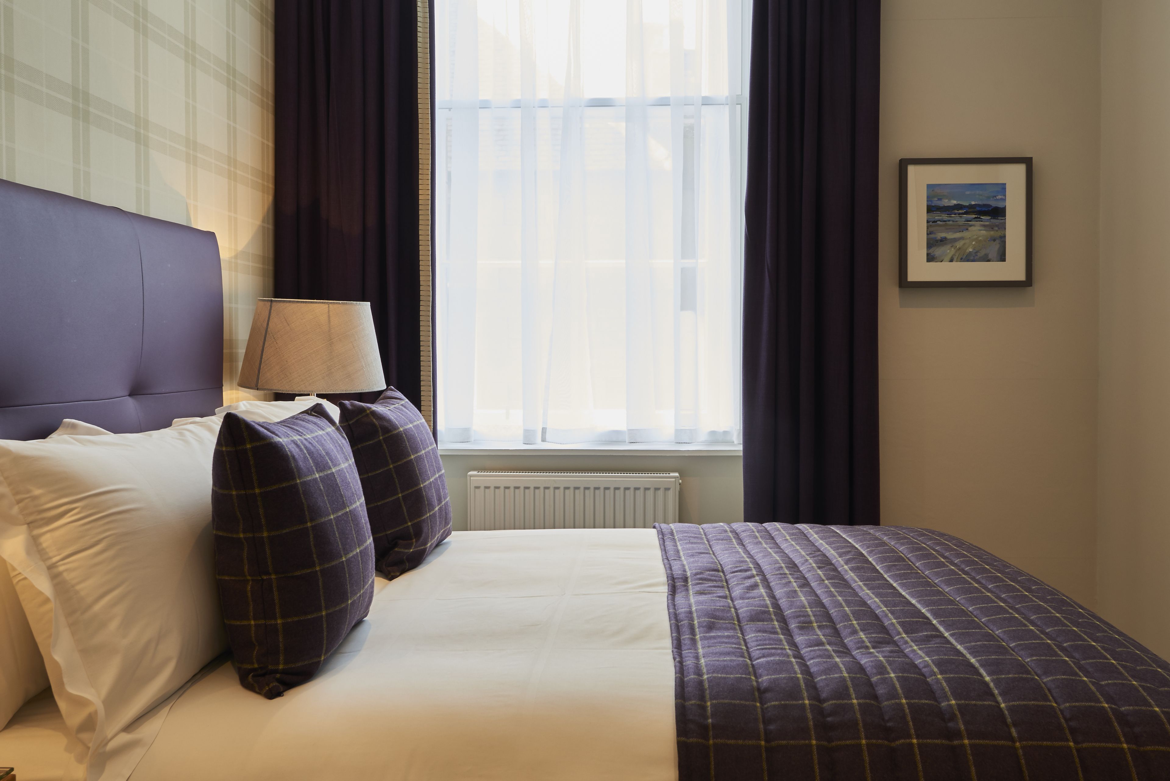 Una cama bien hecha con ropa de cama blanca y morada, una lámpara de noche y una ventana con cortinas.