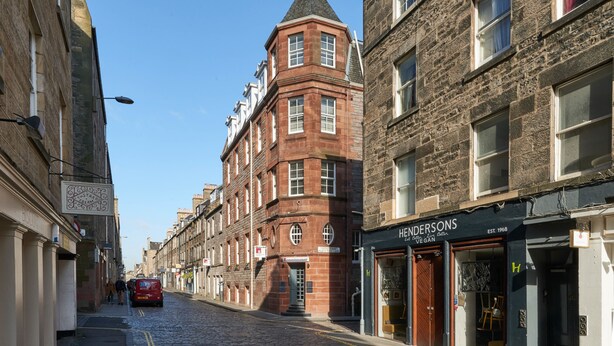 Une rue pavée bordée de bâtiments historiques, dont un immeuble d’angle doté d’une tour remarquable et un magasin dont l’enseigne porte l’inscription « HENDRONS ».