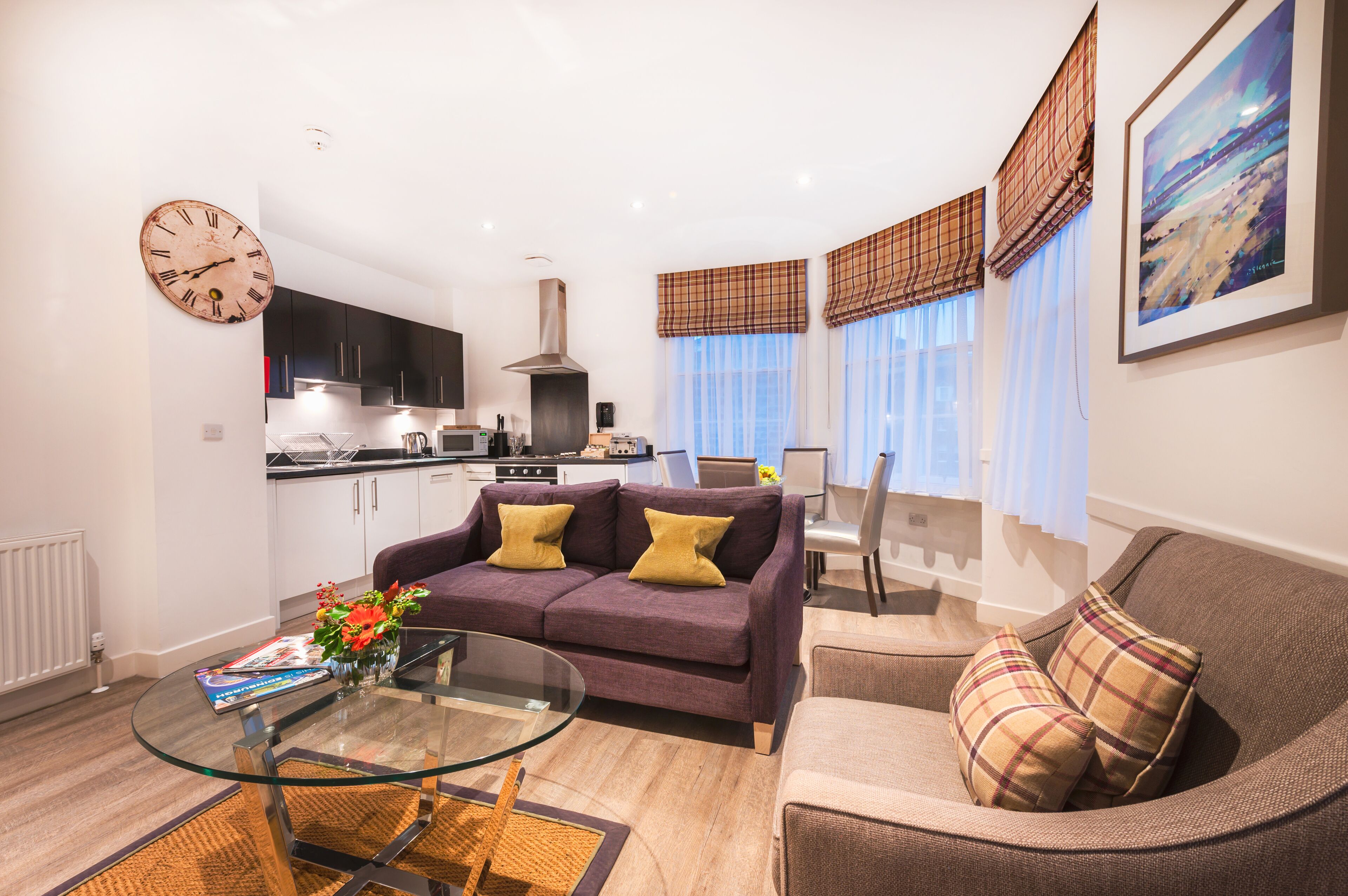 Una sala moderna con sofá, sillones, una mesa de centro de vidrio y una cocina al fondo.