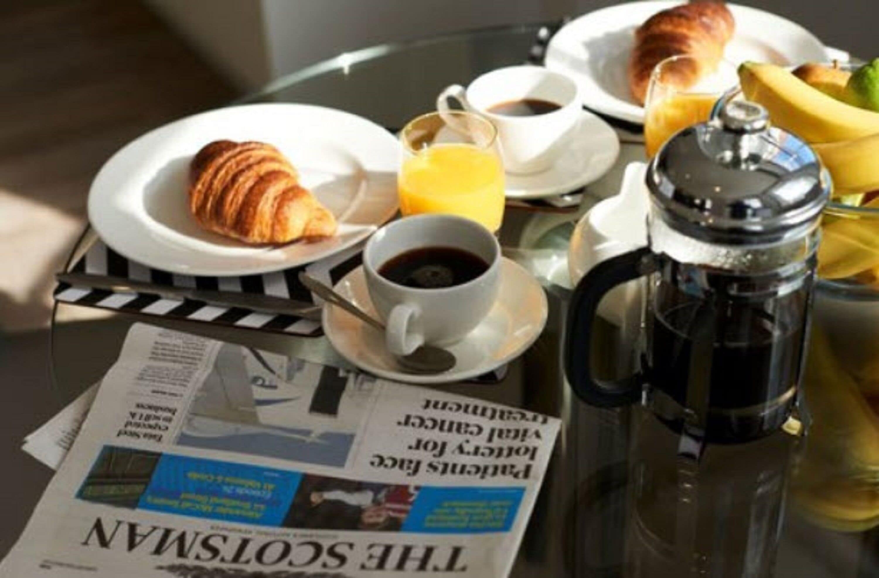 A breakfast setting with croissants, coffee, juice, and a newspaper on a table.