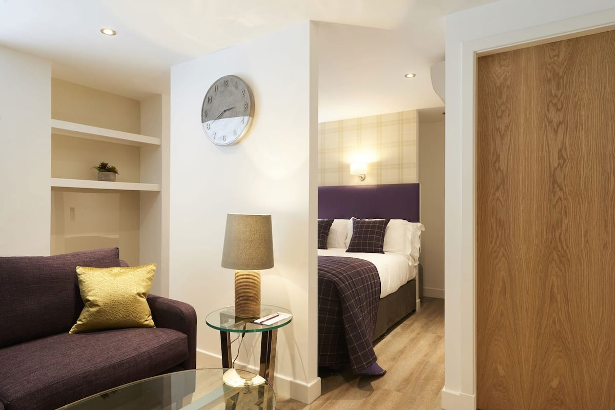 A modern hotel room with a glass coffee table, a sofa with a cushion, a bed with a headboard, and a wall clock.