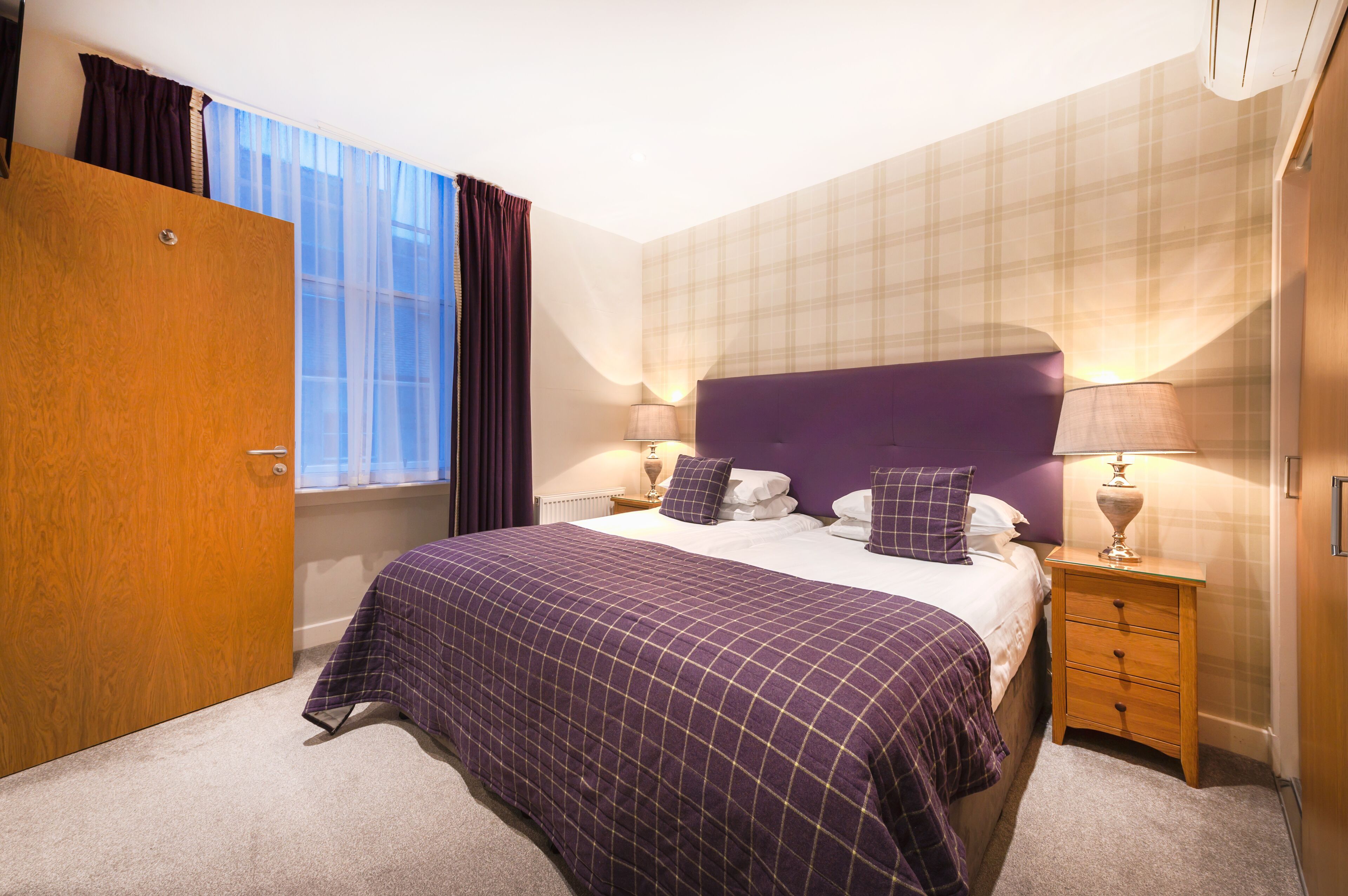 A bedroom with a large bed, a wooden wardrobe, a nightstand with a lamp, and a window with curtains.