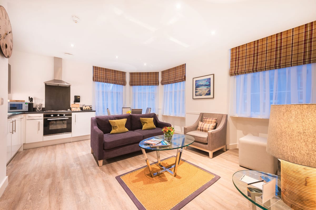 A modern living room with a sofa, armchair, glass coffee table, and a kitchen area with a stove and cabinets.