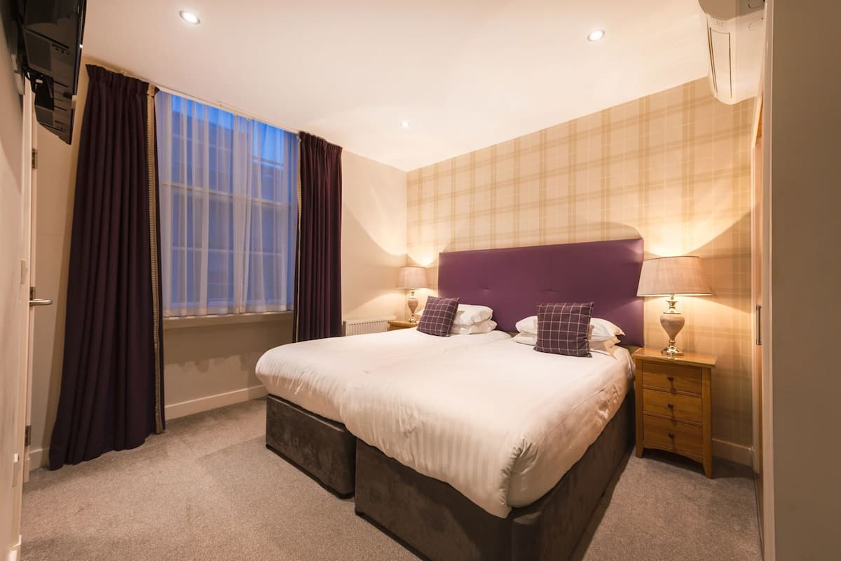 A bedroom with a large bed, two bedside tables, a window with curtains, and a wall-mounted TV.