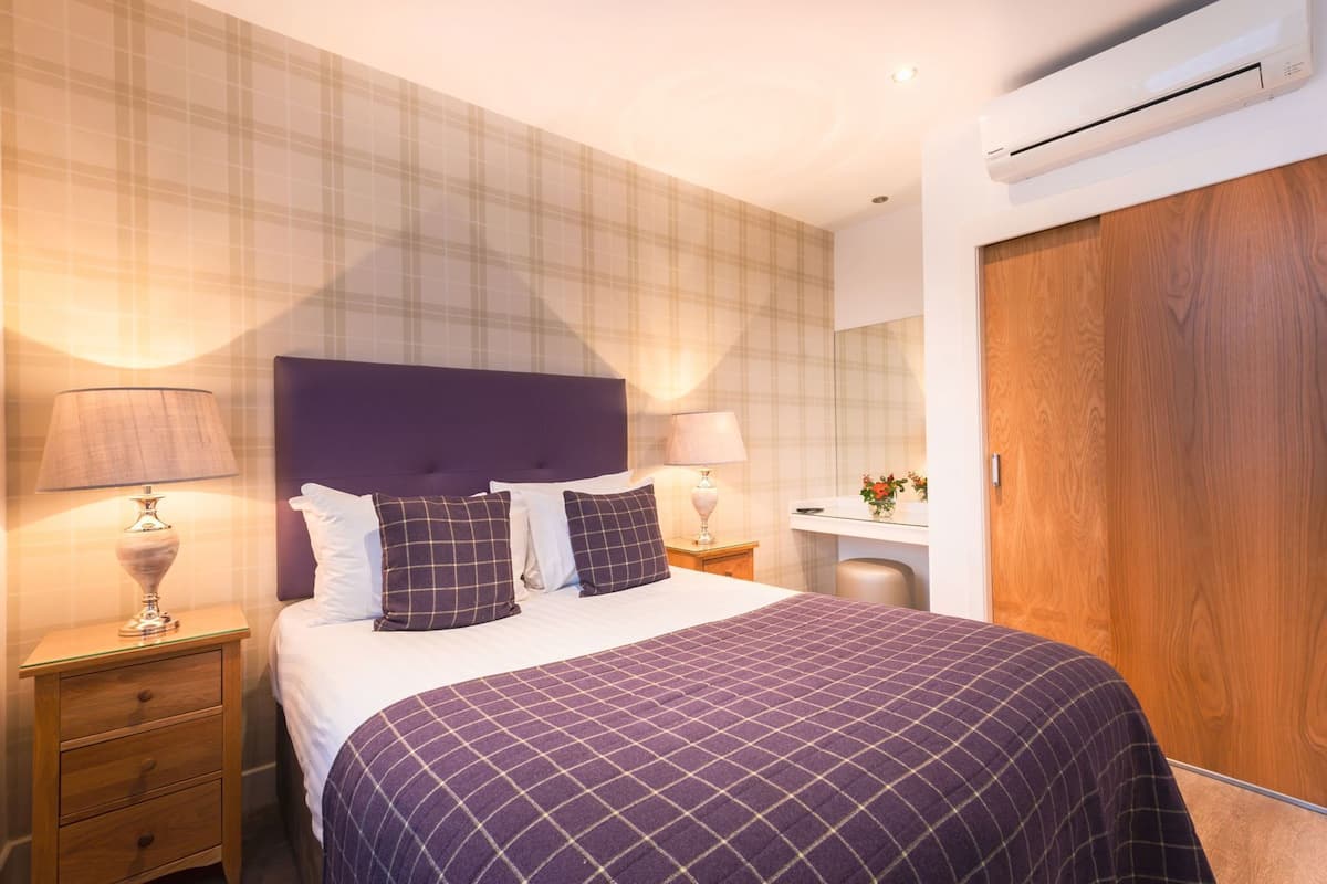 A hotel room with a bed, two bedside tables with lamps, a dresser, a wooden wardrobe, a small desk with a mirror, and an air conditioning unit.