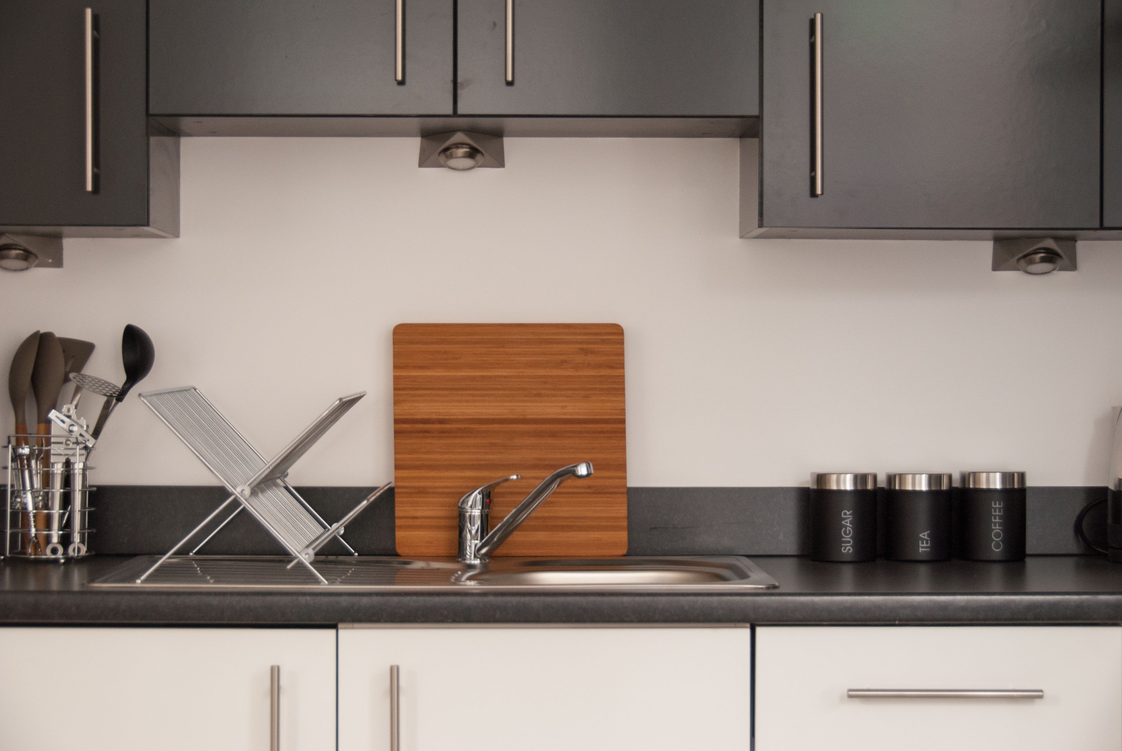 A modern kitchen with a stainless steel sink, a wooden cutting board, and three black containers labeled sugar, tea, and coffee.